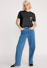 Schwarzes T-Shirt mit kurzen Ärmeln und einer Tasche mit Blumenstickerei, kombiniert mit hoch taillierten, weiten blauen Jeans und schwarzen Schuhen.