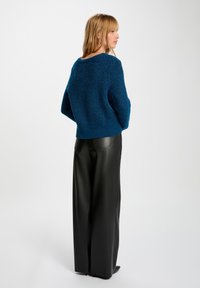 Oversized blauwe gebreide trui met textuurafwerking, ronde halslijn en geribbelde zoom, gecombineerd met wijdlopende zwarte neplederen broek.