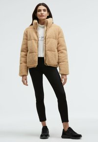 Beige faux fur cropped puffer jacket with a high collar, zip closure, and elastic hem. Paired with black skinny jeans and black sneakers.