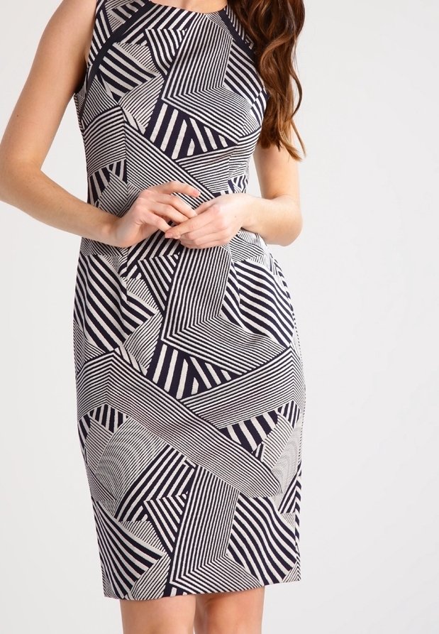 Femme portant une robe sans manches longueur genoux avec un motif rayé diagonal géométrique noir et blanc, mains jointes à la taille.