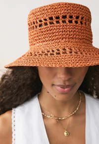 Sombrero de ganchillo en color naranja con detalles cortados en la corona. Presenta un ala ancha, tejido texturizado, combinado con una blusa blanca y collares en capas.