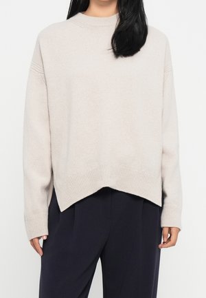 Femme portant un pull côtelé beige clair avec un ourlet en forme de V et des manches longues, associé à un pantalon ample foncé.