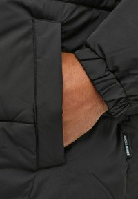 Musta toppatakki, jossa on sivutasku ja joustavat hihansuut, sileä ulkopinta. Alapuoressa näkyvissä brändäystarra.