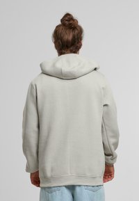 Sudadera de color gris claro, de corte grande, con una gran capucha ajustable. Tejido suave, ajuste casual. Presenta hombros caídos y puños de canalé.