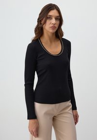 Zwarte ribbelige langemouwen top met een scoopnecklijn, versierd met een metallic kettingdetail. Gecombineerd met lichtbeige broek.