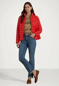 Kvinne iført en rød boblejakke over en mønstret genser, blå oppbrettede jeans og brune loafers, står med den ene hånden i lommen.