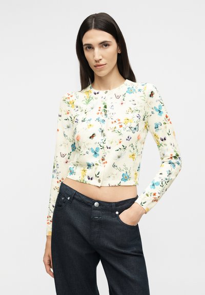 Γυναίκα με μακριά σκούρα μαλλιά που φοράει εφαρμοστή φλοράλ μπλούζα με μακριά μανίκια και παντελόνι με ψηλή μέση σε σκούρο χρώμα, ποζάρει με το ένα χέρι στην τσέπη.