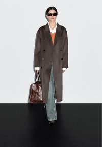 Brązowy płaszcz wełniany z podwójnym zapięciem, pomarańczowy sweter pod białą koszulą z kołnierzykiem, jasnoniebieskie dzwony, brązowa skórzana torebka oraz czarne szpilki.