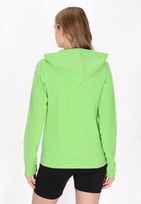 Giacca in pile verde chiaro con cappuccio, maniche lunghe e una texture morbida. Pantaloni corti neri visibili in basso. Design semplice e funzionale.