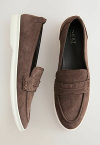 Mocassins marron en daim à enfiler avec semelles en caoutchouc blanches, ornés d'une lanière cousue sur le dessus.
