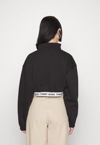 Černá croppovaná mikina s vysokým límcem a oversized rukávy, s bílým páskem s logem. Béžové kalhoty se zadními kapsami.