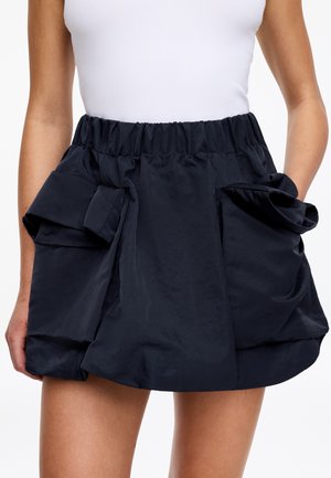 Donna che indossa una gonna corta nera e voluminosa con tasche arricciate e vita elastica, abbinata a una canotta bianca senza maniche.