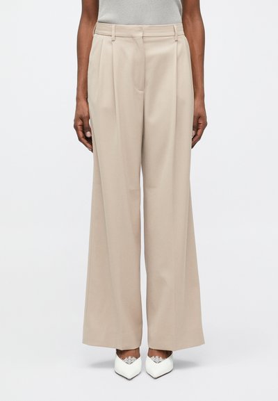 Pantalon beige à jambes larges avec plis, en tissu lisse ; assorti de chaussures blanches à bout pointu avec des détails en dentelle.