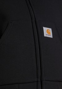 Felpa nera con zip, tasche frontali e patch quadrato bianco con logo Carhartt che presenta un simbolo arancione e il nome del marchio sulla tasca destra.