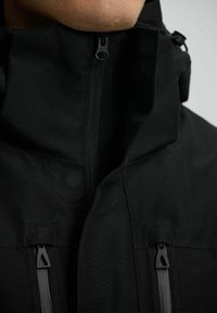 Giacca nera impermeabile con alto colletto, due tasche con zip e una texture liscia. La cerniera YKK presenta un design minimalista.