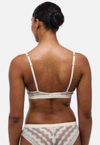 Soutien-gorge en dentelle beige clair avec des bretelles réglables, des bords festonnés et un dos texturé. Culotte en dentelle assortie visible.