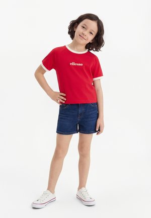 Παιδί με κοντά σκούρα μαλλιά, ντυμένο με κόκκινη μπλούζα ellesse, τζιν σορτς και λευκά αθλητικά παπούτσια, στέκεται με το ένα χέρι στη μέση.