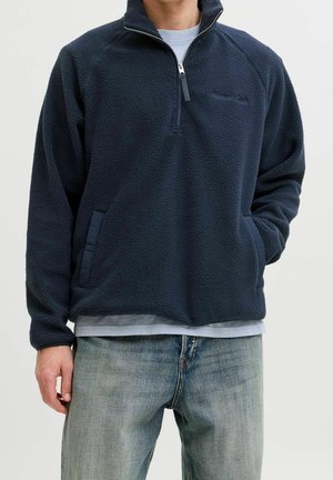 Homme portant un pull polaire bleu marine à zip quart avec poches avant et un jean bleu clair sur un fond uni.