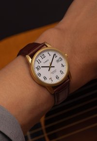 La montre-bracelet Timex présente un cadran blanc, des chiffres arabes noirs, un boîtier doré et un bracelet en cuir brun à texture crocodile. Affichage de la date inclus.