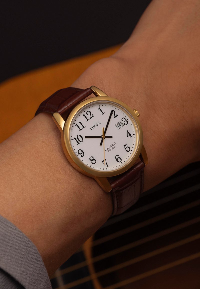 La montre-bracelet Timex présente un cadran blanc, des chiffres arabes noirs, un boîtier doré et un bracelet en cuir brun à texture crocodile. Affichage de la date inclus.
