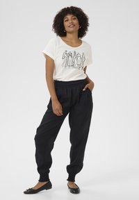 T-shirt bianco con grafica nera di sei figure in abito, abbinato a pantaloni jogger neri con vita elasticizzata e ballerine nere.