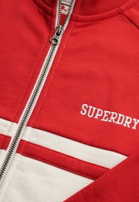 Rode jas met ritssluiting gemaakt van zacht materiaal, met witte horizontale strepen en "SUPERDRY" in het wit geborduurd. Zilveren ritssluiting met trekkoordje.