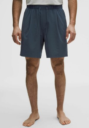 Man draagt donkerblauwe elastische short met trekkoorden, staat op blote voeten tegen een effen lichte achtergrond.