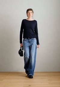 Črn dolg rokav majica in svetlo modri široki kavbojke. Model drži majhno črno torbico. Gladek lesen pod je in enostavna siva ozadje.