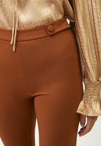 Pantaloni marroni aderenti con una texture liscia, caratterizzati da un dettaglio con bottone in vita e abbinati a una blusa dorata plissettata.