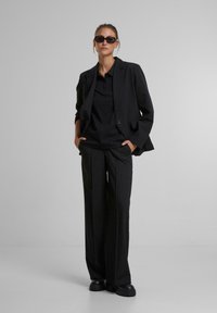 Černý blazer přes černé polo, v kombinaci s volnými černými kalhotami a robustními černými botami. Má elegantní, minimalistický design a dobře padnoucí střih.