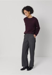 Bordový svetr s dlouhými rukávy, v kombinaci se šedými kalhotami na míru. Modelka má na sobě černé kotníkové boty, ruce v kapsách, neutrální pozadí.