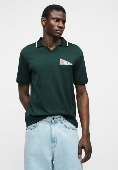 Mężczyzna w ciemnozielonej koszulce polo z krótkim rękawem i naszywką "FA Club" oraz jasnobłękitnych jeansach, stojący z rękami w kieszeniach na białym tle.