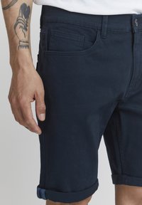 Sötétkék pamut rövidnadrág, slim fit fazonban, felhajtott szegélyekkel, oldalsó zsebekkel és diszkrét varrási részletekkel, egy tetoválással bíró kézmódszerű modellen viselve.
