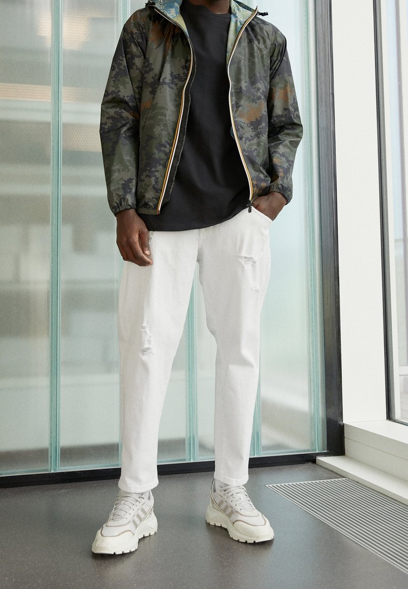 Homme portant une veste camouflage, un t-shirt noir, un pantalon blanc déchiré et des baskets blanches, debout à l'intérieur près de panneaux en verre et d'une bouche d'aération au sol.