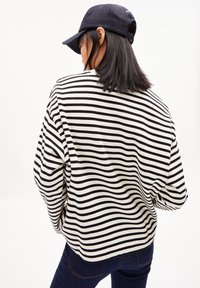 Langärmliges gestreiftes Hemd mit schwarz-weißen horizontalen Linien, das einen lockeren Schnitt und weichen Stoff hat. Getragen mit dunklen Jeans und einer Kappe.