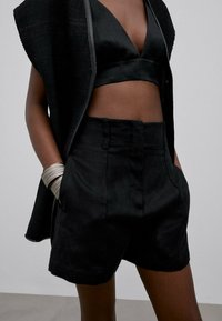 Un haut court noir et un short ajusté en tissu texturé, associés à une couche extérieure en maille ouverte noire et un bracelet à motifs.