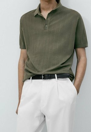 Man draagt een olijfgroen textuur polohemd met korte mouwen, ingestopt in witte geplooide broek met een zwarte riem, één hand in de zak.