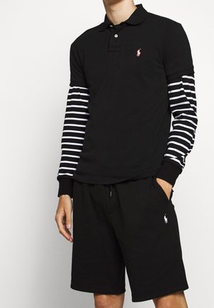 Man draagt een zwarte polo met witte en zwarte gestreepte lange mouwen eronder, gecombineerd met een zwarte korte broek, staat met één hand in de zak.