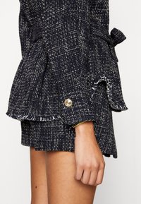 Blazer en tweed bleu marine avec un motif texturé, ourlet à franges et boutons dorés en guise d'accent. Se noue à la taille et présente une coupe ajustée.