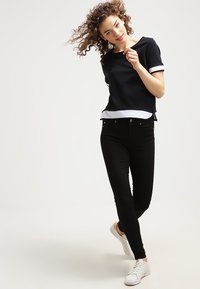 Top corto nero con rifiniture bianche, abbinato a jeans neri aderenti e scarpe da ginnastica bianche, con un design elegante e semplice.