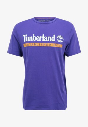 Fialové bavlněné tričko s krátkým rukávem, které má bílé logo Timberland a nápis "ESTABLISHED 1973" v oranžové barvě přes hruď.