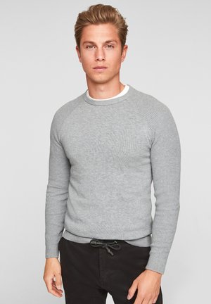 Suéter gris de punto con textura acanalada, cuello redondo y mangas largas. Lleva puesto un pantalón negro con cordón. Diseño sencillo, sin patrones ni acentos.