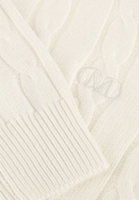 Närbild av en krämfärgad kabelstickad tröjsleeve med ribbad mudd och subtilt broderad monogram på den strukturerade tyget.