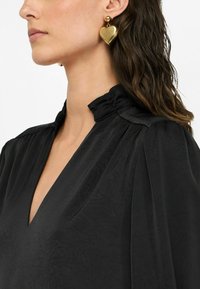 Blusa nera con scollatura a V profonda e un dettaglio della collana arricciato. Presenta grandi orecchini a forma di cuore dorati con una superficie liscia e riflettente.