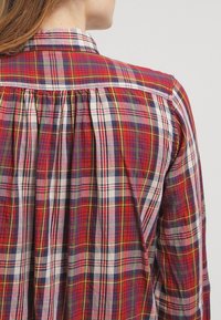 Camicia a quadri rossa e blu in morbido tessuto, con colletto classico e dettaglio arricciato sulla schiena. Accenti bianchi lungo le cuciture.