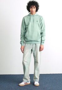 Mintgroene hooded sweatshirt met een voorzak en ribbels aan de manchetten, gecombineerd met lichtgewicht, licht versleten jeans en witte sneakers met rode accenten.