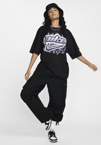 Černé oversized grafické tričko s fialovým logem Nike a designem plamenů, doplněné černými cargo kalhotami a černobílými teniskami.