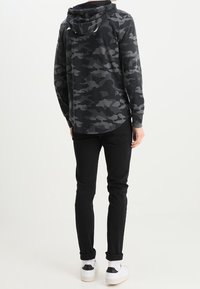 Svart camouflagetröja med lång ärm och huva, bärs med svarta byxor och vita sneakers. Enkelt design, mjukt tyg och avslappnad passform.