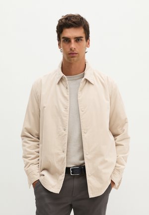 Junger Mann mit dunklen Haaren trägt eine beige Jacke mit Knöpfen über einem hellen Pullover, dunkle Hose und schwarzen Gürtel, steht vor einem schlichten weißen Hintergrund.