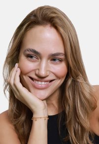 Bracciale d'oro con ciondoli a goccia appesi a un polso, indossato da una donna con lunghi capelli castano chiaro e ondulati e un sorriso dolce.